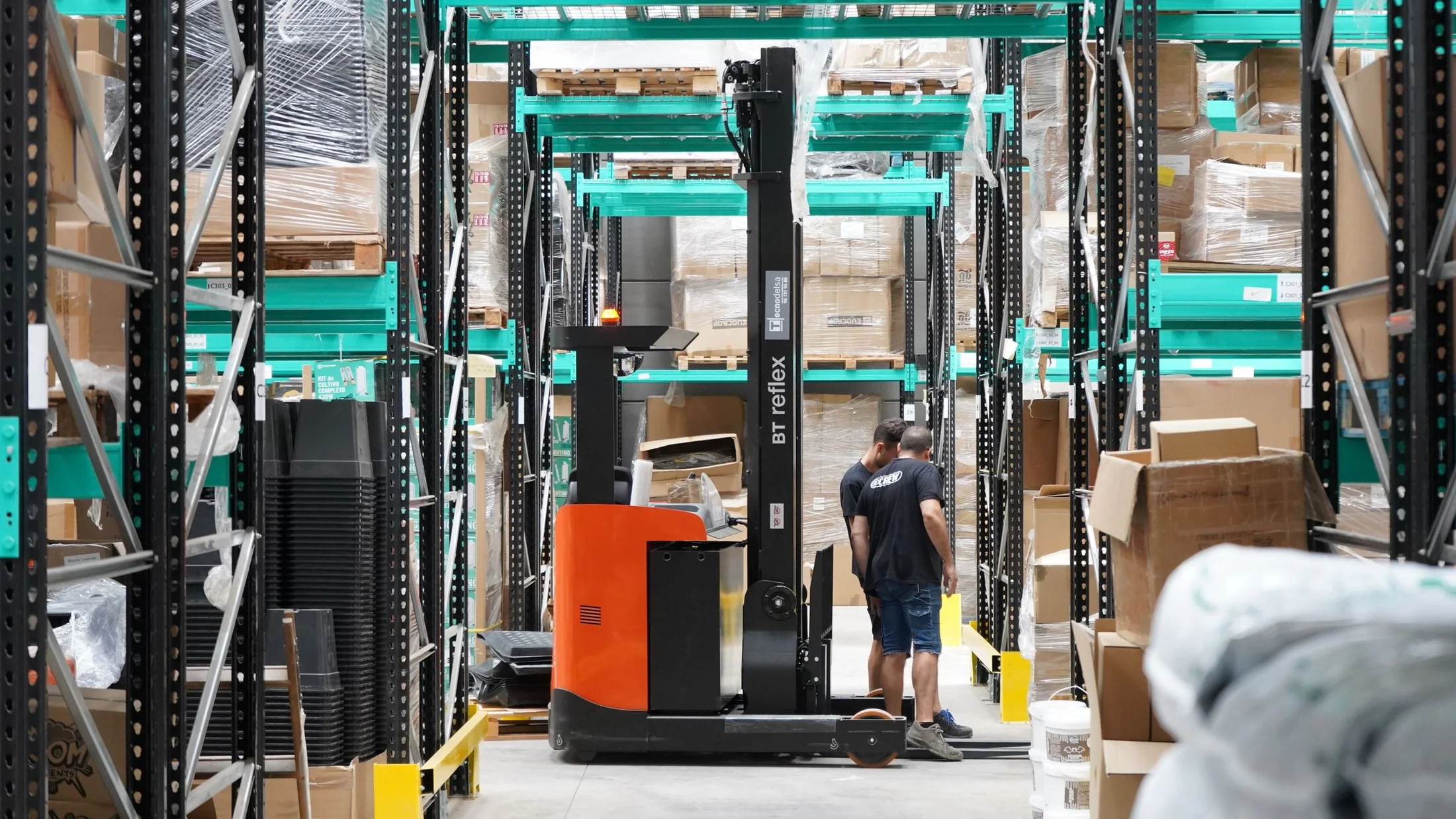 Two warehouse workers beside material-handling equipment in a stocked aisle